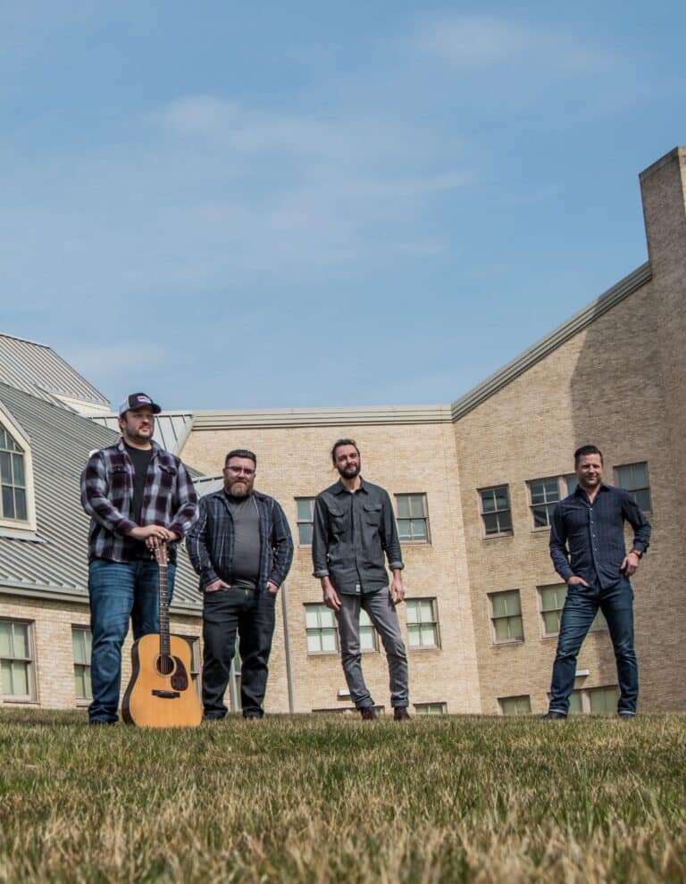 Four men standing on a grass lawn with an acoustic guitar, posed in front of a brick building