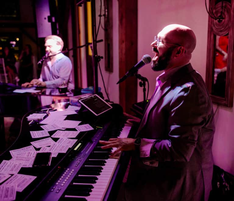 Two pianists singing and playing dueling pianos under purple stage lights at Founders Hall