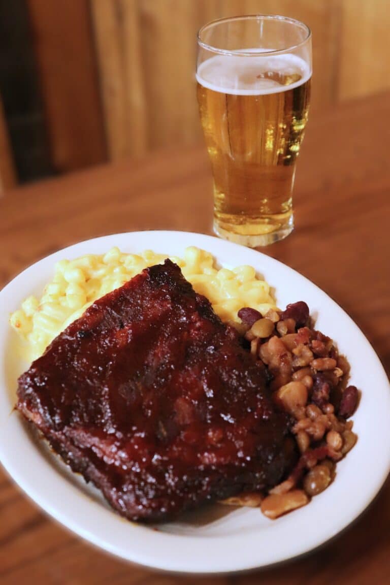 BBQ ribs with mac and cheese and baked beans beside a glass of beer on a wood table at the Barn Restaurant