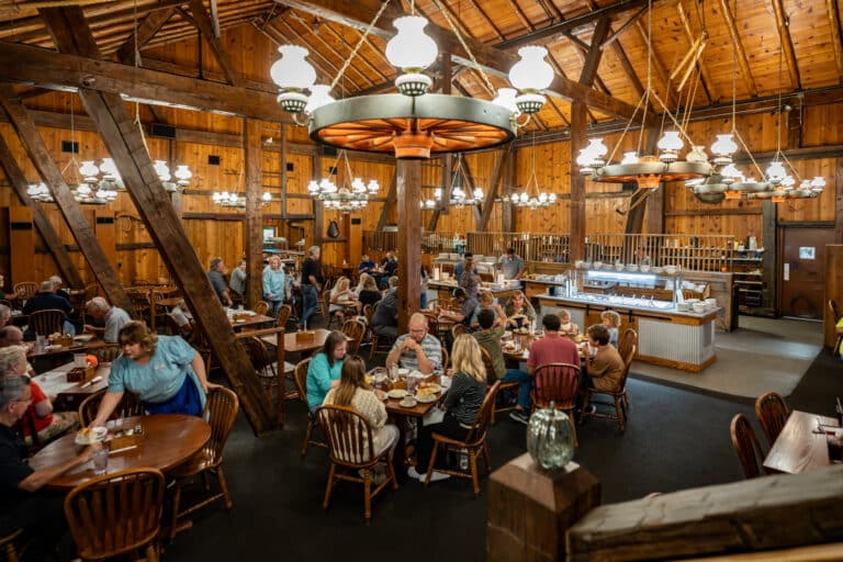 Guests dining inside the Barn Restaurant at Sauder Village, beneath wagon-wheel chandeliers and warm wood-beamed ceilings