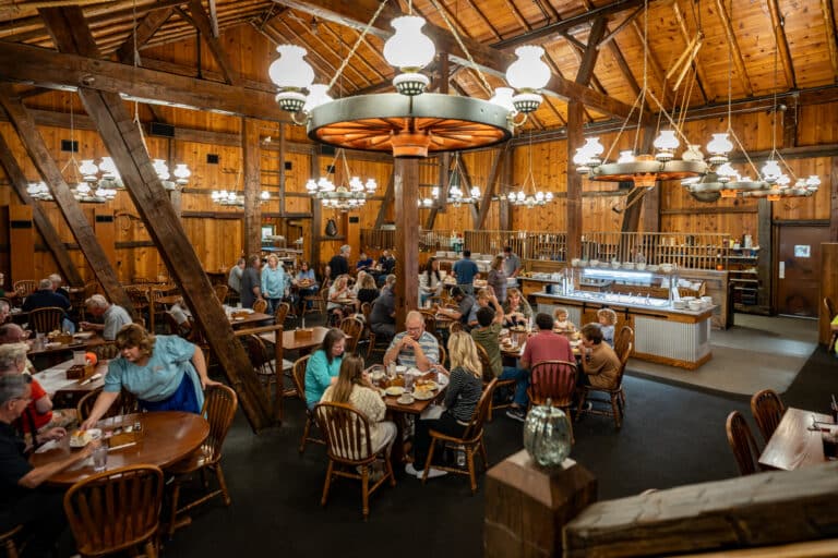 Families enjoying a meal inside the Barn Restaurant at Sauder Village, with rustic wood walls and hanging lantern chandeliers