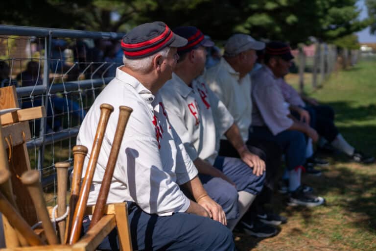 Baseball players in vintage uniforms sit on a bench with wooden bats during a living-history game