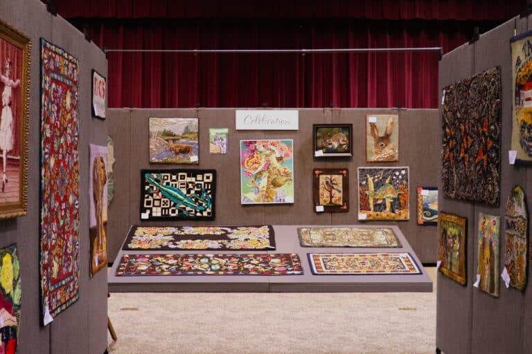 Hooked rug display booth labeled "Celebration" featuring animal portraits, floral rugs, and patterned pieces at a Sauder Village fiber arts show
