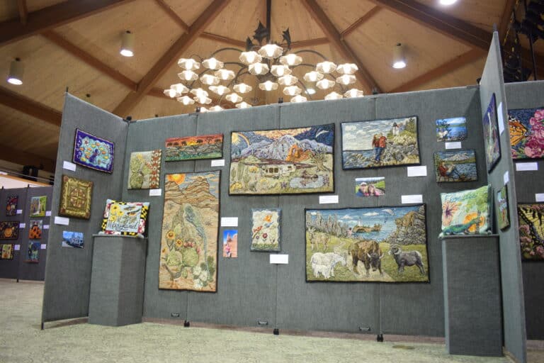 Colorful hooked-rug artwork displayed on gray panels inside a wood-beamed hall at a Sauder Village maker retreat