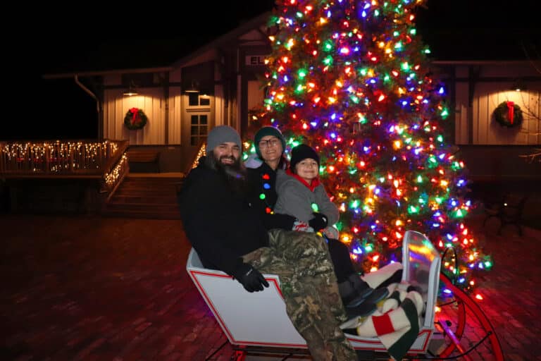 Family of three smiling in a holiday sleigh beside a lit Christmas tree at Sauder Village after dark