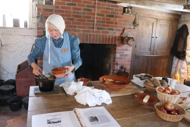 Costumed interpreter in bonnet and apron cooking over an open hearth in a historic kitchen