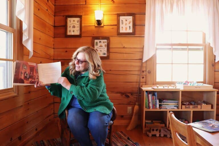 Woman in green jacket reads a picture book aloud in a cozy wood-paneled room at Sauder Village