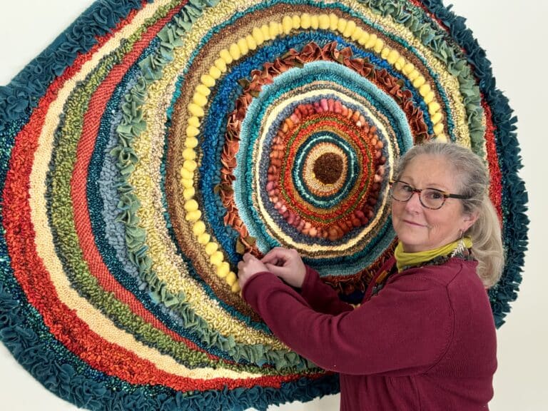 An older woman with gray hair and glasses smiles beside a large circular fiber art piece mounted on a wall