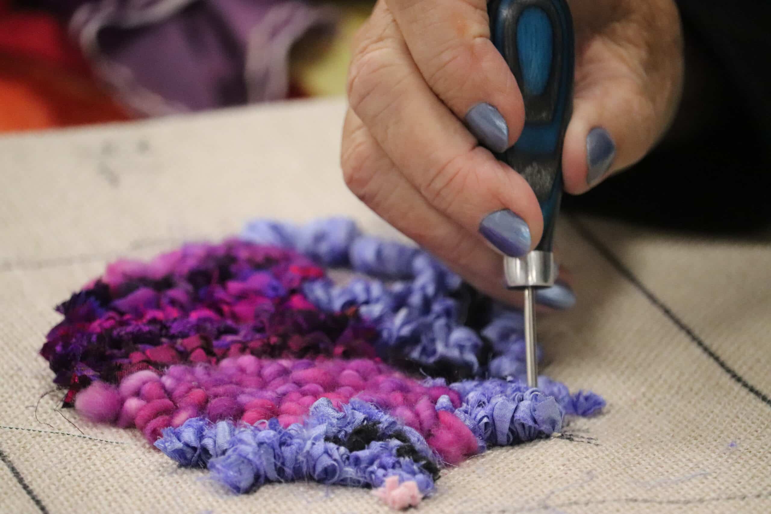 Close-up of a punch needle pushing colorful purple and pink fabric strips through linen backing during a rug hooking class