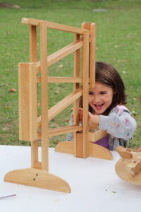 A young girl laughs while playing with a wooden marble run at an outdoor table