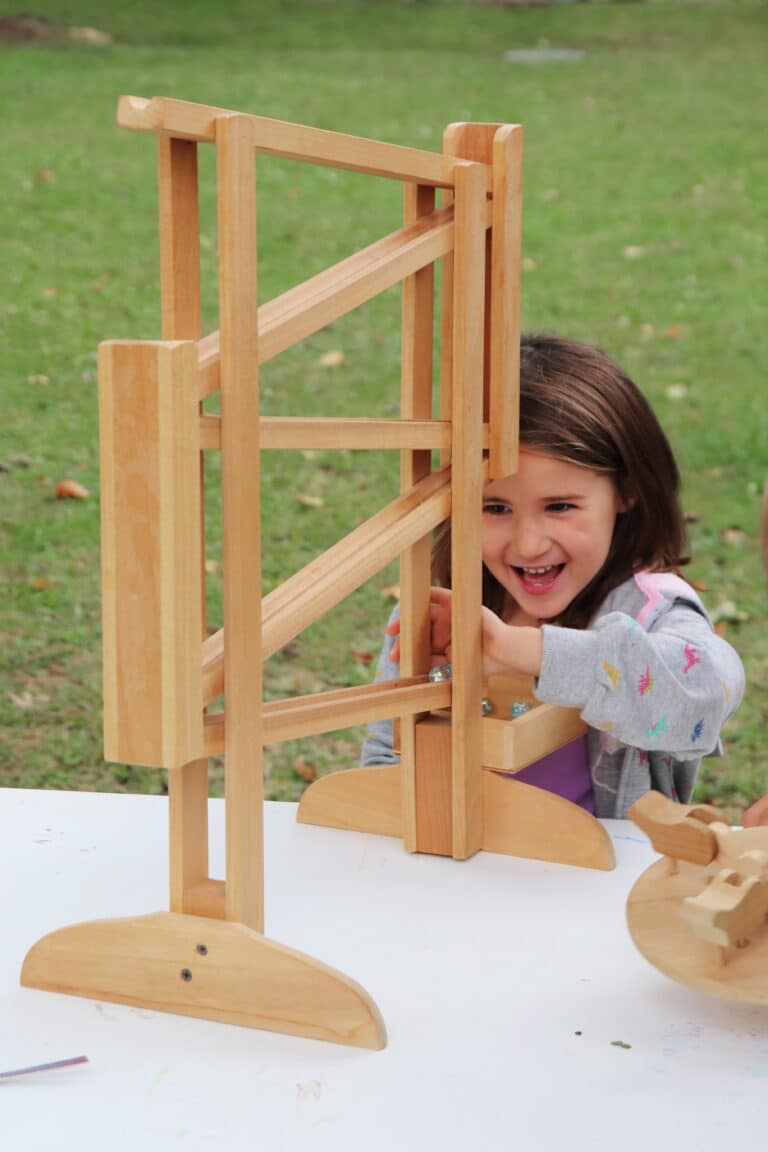 A young girl laughs while playing with a wooden marble run at an outdoor table