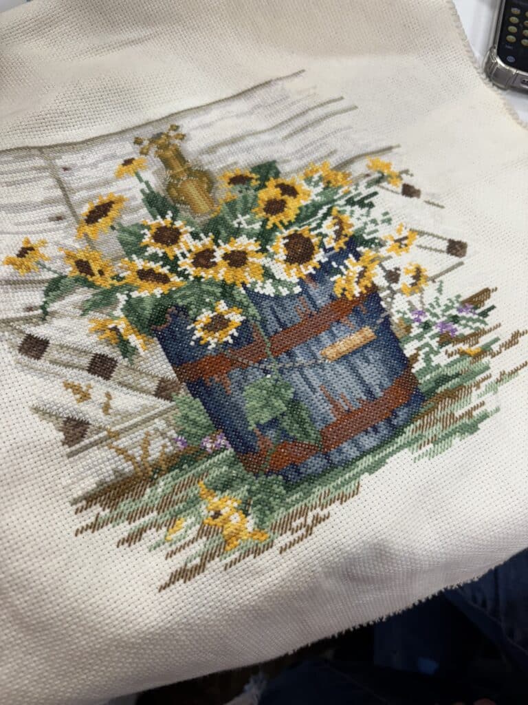 Detailed cross-stitch of sunflowers in a blue wagon, in progress on cream Aida cloth