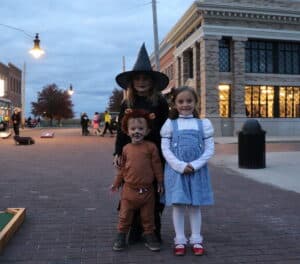 Three children in Wizard of Oz Halloween costumes trick-or-treating on a brick street at dusk