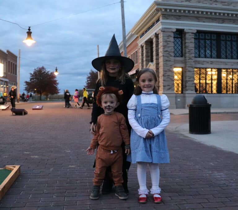 Three children in Wizard of Oz Halloween costumes trick-or-treating on a brick street at dusk