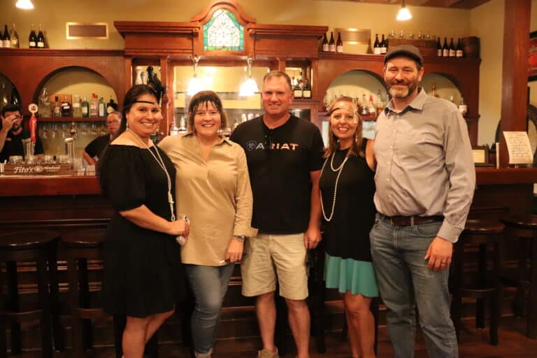 Five smiling guests pose together in front of a classic wooden bar at a Sauder Village evening event