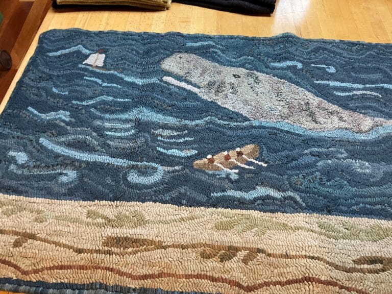 Hand-hooked rug depicting a gray whale and small rowboat on a blue ocean with a sandy shore in the foreground