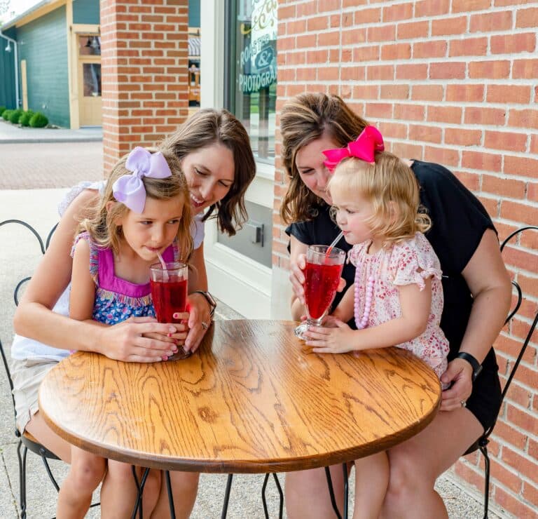 Two moms and their young daughters sip red drinks at an outdoor café table beside a brick storefront