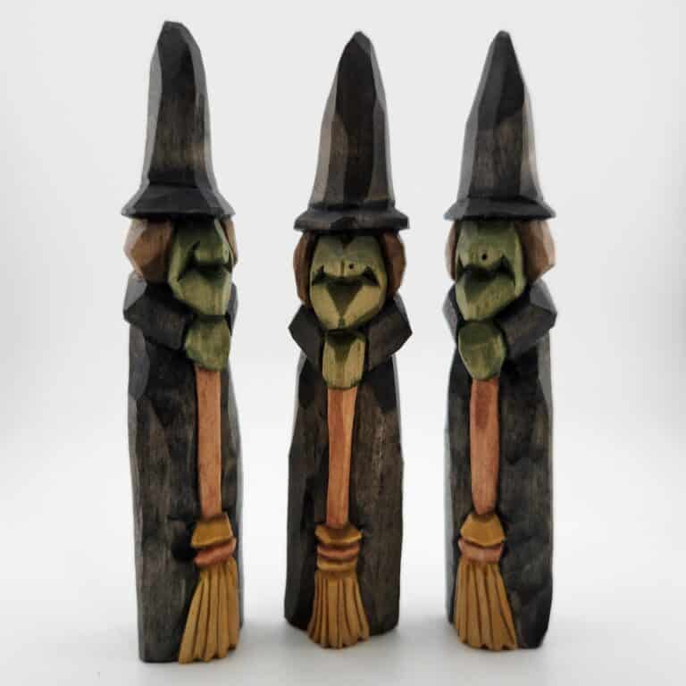 Three dark-stained wood-carved witch figures holding brooms, with green faces and pointed black hats