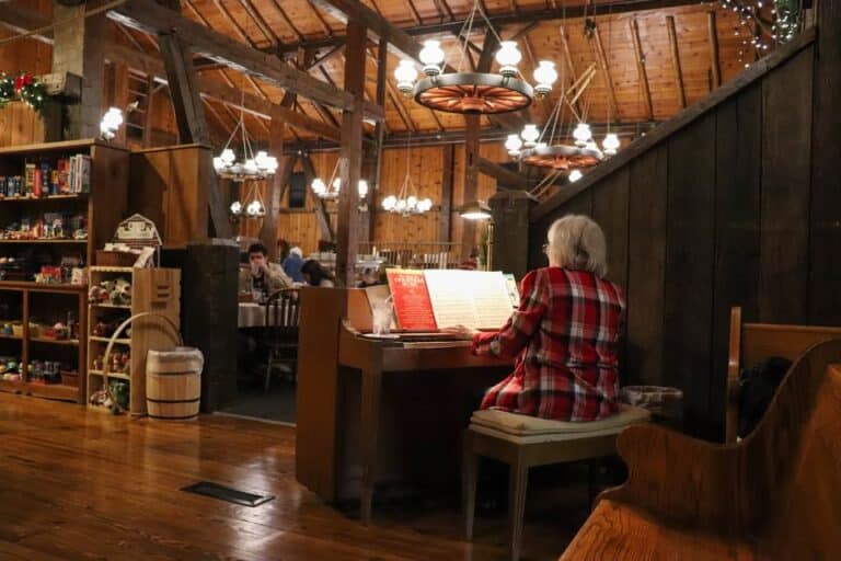 Elderly woman playing piano in the rustic, warmly lit Barn at Sauder Village, with wagon-wheel chandeliers overhead