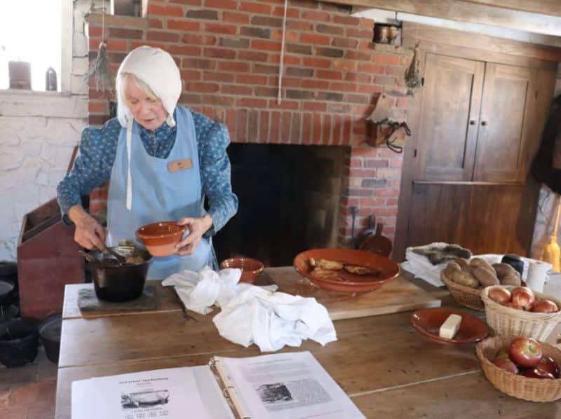 Costumed interpreter preparing food beside a brick hearth